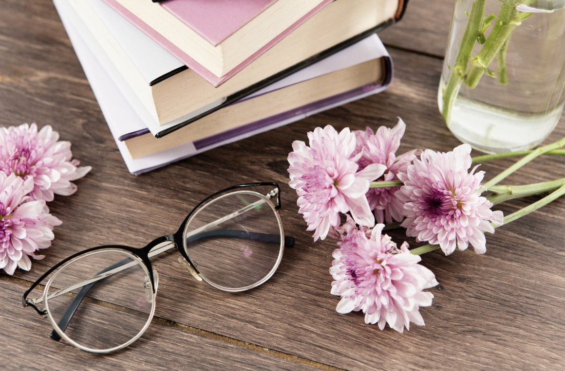 Black-rimmed eyeglasses with clear lenses on wooden table beside pink flowers and stacked books