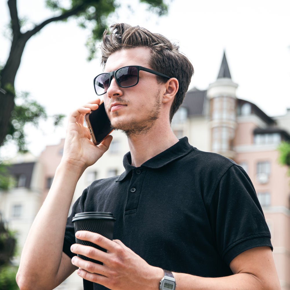 Man wearing stylish sunglasses while talking on phone and holding coffee outdoors
